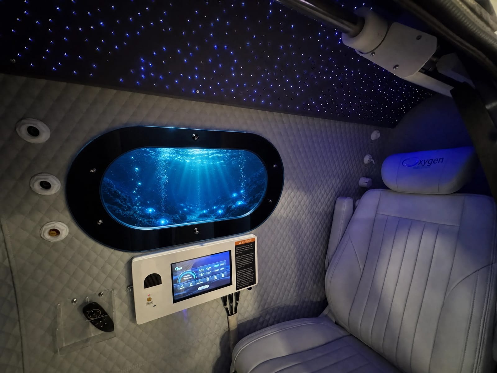 Interior of the Executive XLS-35 with the starlight fiber-optic ceiling glowing above the quilted walls, the oval window showing an underwater ocean scene, the touchscreen control panel, and the premium reclining seat